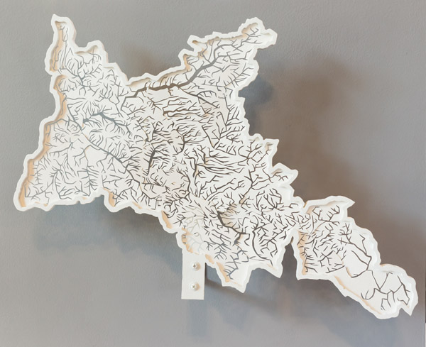 A white sculpture showing the streams of a watershed. The sculpture is shaped like a watershed boundary. Most of the sculpture is flat, but its boundary is raised. There are many cuts in the sculpture, though which a grey wall can be seen. The cuts form the shape of a stream network. Photo by Charles P. Rader.