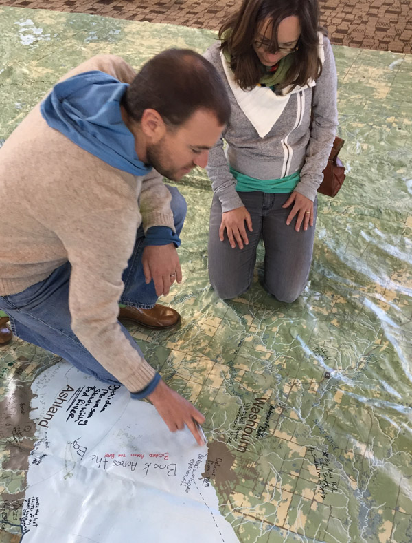 Two people kneeling on top of the large map. One is pointing to the map, appearing to bring it to the attention of the other person. Photo by Matt Dooley.