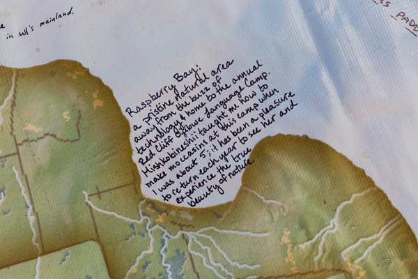 A close-in photograph of part of a map. Upon the map is written, in black marker, 'Raspberry Bay: a pristine natural area away from the buzz of technology & home to the annual Red Cliff Ojibwe Language Camp. Mishkobineshii taught me how to make moccasins at this camp when I was about 5; it has been a pleasure to return each year to see her and experience the true beauty of nature.' Photo by Charles P. Rader.
