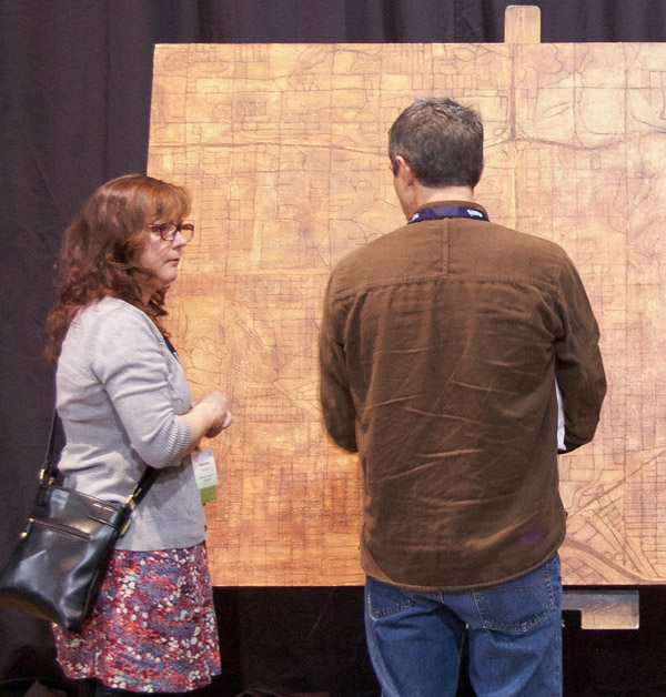 Two people standing in front of the brown map. They appear engaged in conversation. The map is mostly square and several feet wide. Photo by Dylan Moriarty.