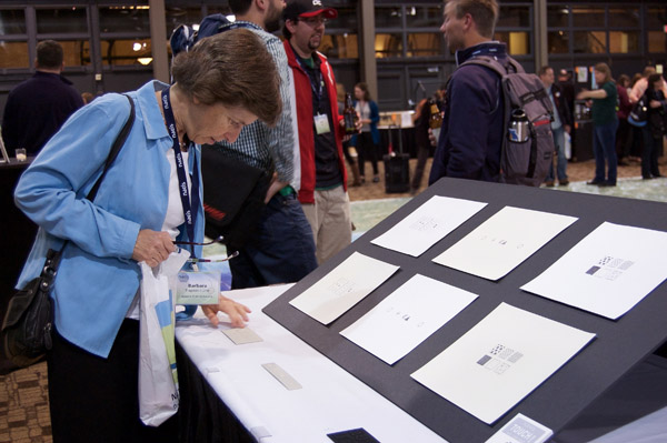 A person standing in front of a table. On the table is a large piece of black board, on which are several sheets of white paper. The person is looking down at the table and examining small objects positioned just in front of the black board. Photo by Dylan Moriarty.