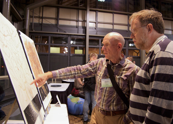 Two people looking at the pair of maps. The maps are on display stands atop a table. One person points to a detail on one of the maps. Photo by Dylan Moriarty.