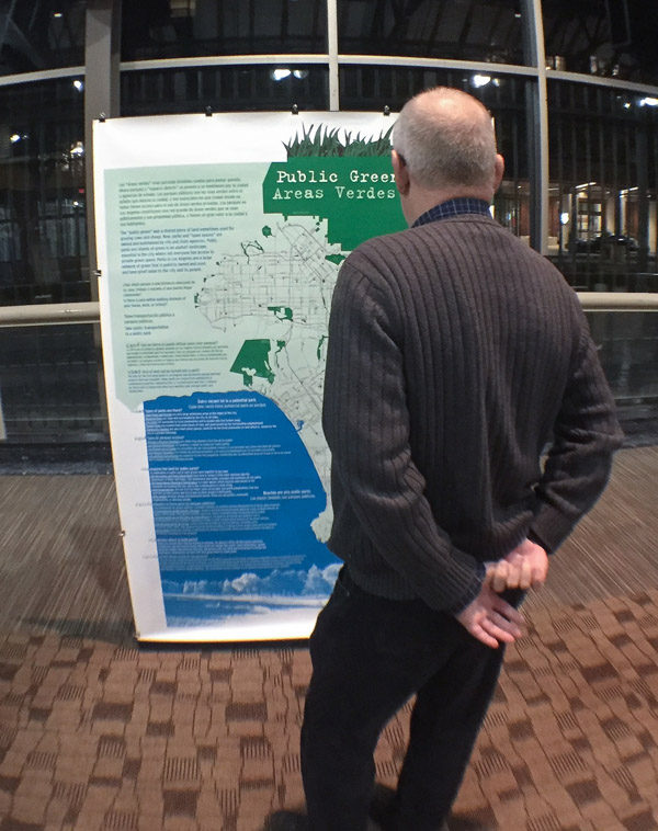 A person, seen from behind, looking at a tall poster standing vertically on a floor. The poster shows a map of green spaces and red roads. Photo by Matt Dooley.