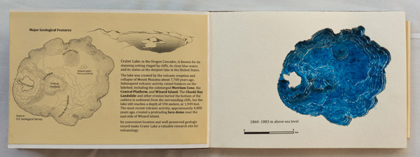 A small book, opened. On the left-hand page, there is a block of text and a map of the depth contours of a lake. On the right, the block of pages have each had holes cut in them to form a three dimensional sculpture of the lake. The sculpture of the lake is painted blue. Photo by Charles P. Rader.