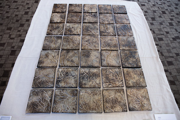A series of 35 similar-looking tiles sitting in an arrangement 5 tiles wide and 7 tiles tall. They are sitting on a white cloth placed on a carpet. Each tile is textured. The texture appears to form a stream network. The tiles are a mottled brown color. Photo by Charles P. Rader.